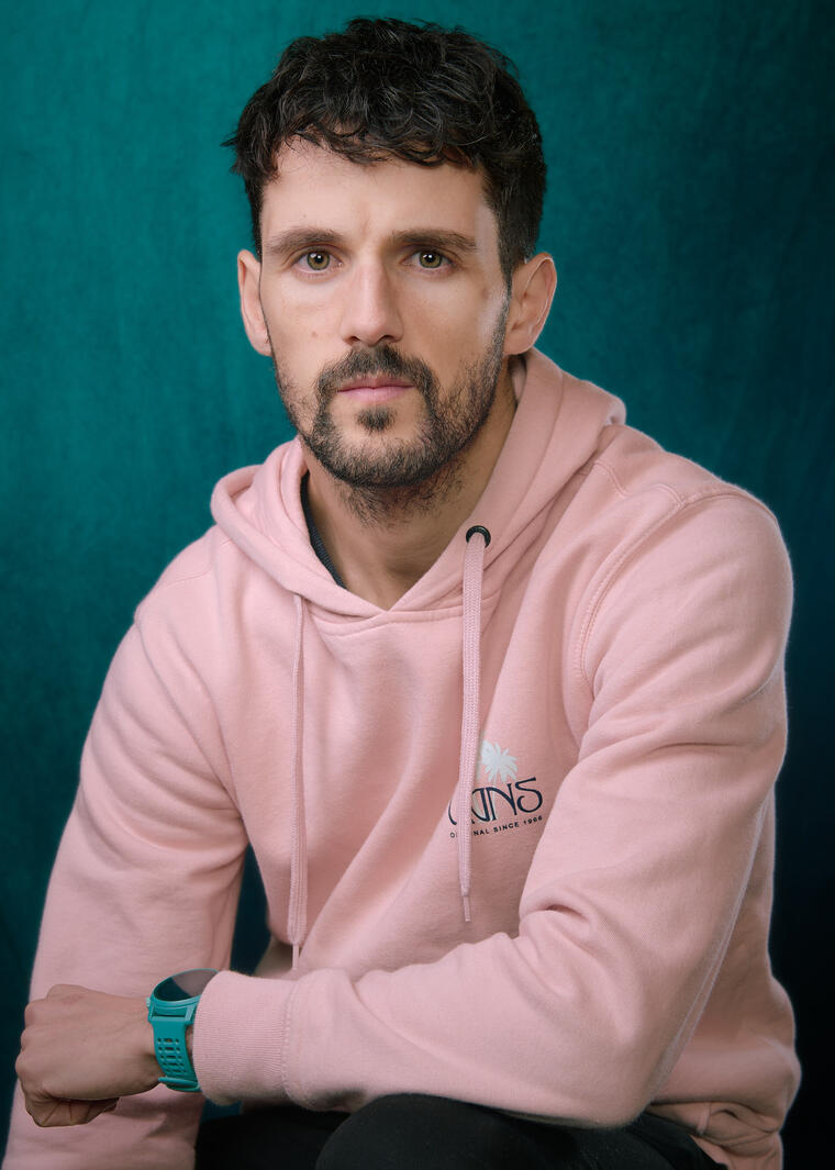 A picture of a 30 year old male wearing a pink jumper with a green background behind him